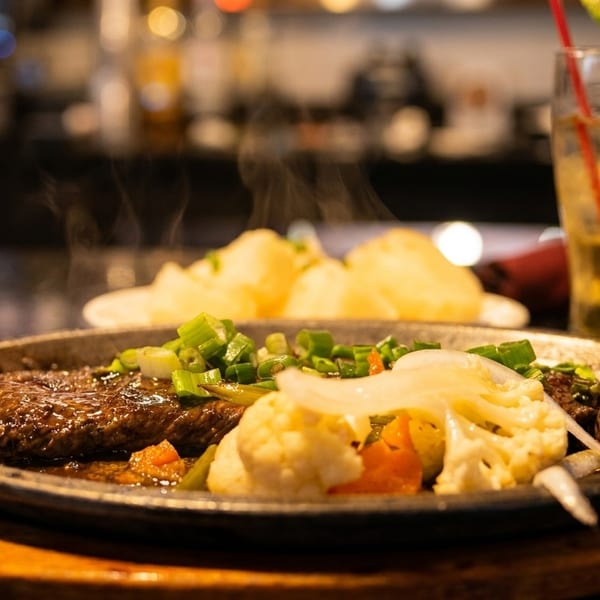 Sizzling steak with vegetables and mashed potatoes, with a drink in the background.