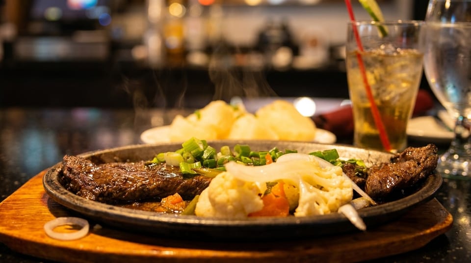 Sizzling steak with vegetables and mashed potatoes, with a drink in the background.