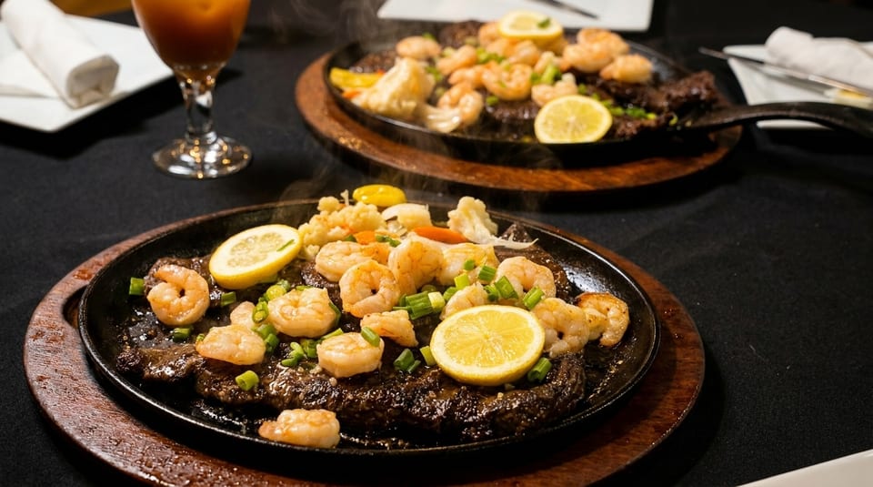 Two sizzling platters of steak and shrimp with lemon slices and chopped green onions.