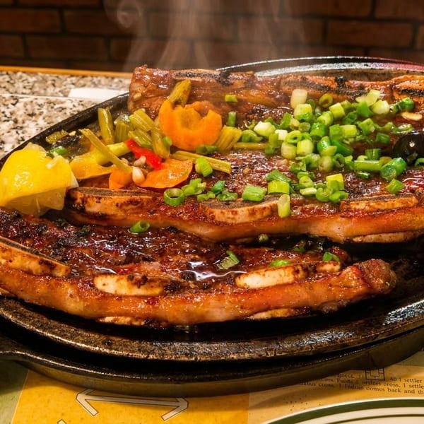 Sizzling grilled short ribs with green onions, vegetables, and lemon on a hot plate.